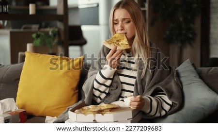 Portrait of sad young blond woman dealing with stress by eating food sitting on sofa at home Upset young woman wrapped in blanket crying and eating pizza indoors Royalty-Free Stock Photo #2277209683