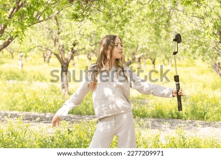 Young girl dancing takes pictures of herself on front camera of smartphone. Park, spring garden with grass, flowering trees. Concept blogging, selfie, spring, childhood, content creation. Caucasian.