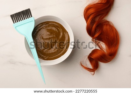 Bowl of henna cream, brush and red strand on white marble table, flat lay. Natural hair coloring