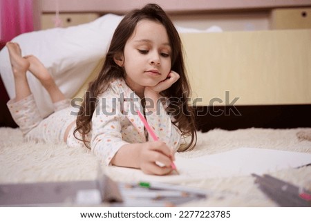 Caucasian little child, a baby girl in stylish pajamas with multicolored dots, drawing picture with colorful pencils indoors in her bedroom. Art. Creativity. Children. Lifestyles. Leisure. Hobby