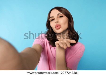 Beautiful young woman taking selfie while blowing kiss on light blue background