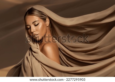 Beautiful Woman wrapped in Beige Silk Scarf. Beauty Girl Face with Smooth Skin and Natural Makeup. Women Hair Sun Care. Headwear Scarves over Desert Sand background Royalty-Free Stock Photo #2277295903
