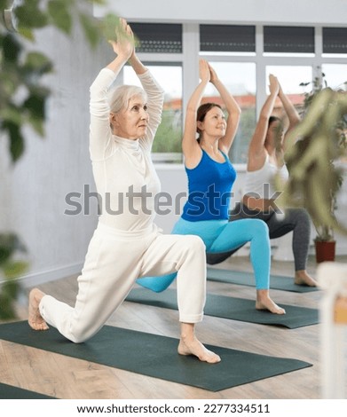Positive mature female stretching in Virabhadrasana 1 or warrior 1 position during group yoga workout in gym studio Royalty-Free Stock Photo #2277334511