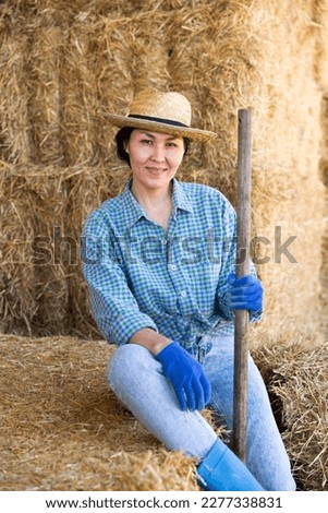 Positive relaxed asian female farmer resting on hayloft after work at farm, sitting on straw stack and smiling