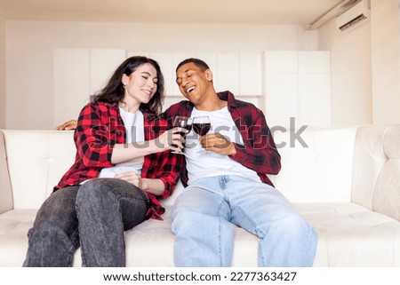 young multiracial couple sitting at home on comfortable sofa talking and drinking wine, african american man with woman on couch clink glasses and smile, interracial friends celebrate event together