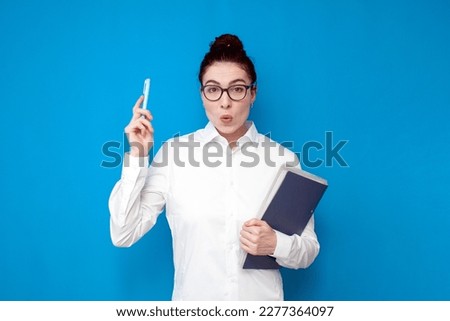 young shocked girl office worker in white shirt and glasses shows surprise with her hand up on blue background, surprised female teacher with documents has idea Royalty-Free Stock Photo #2277364097