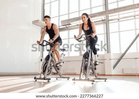 beautiful athletic couple of cyclists train in the fitness room on static bicycle simulator, man trainer and fitness woman go in for sports together on simulators in the morning in bright room