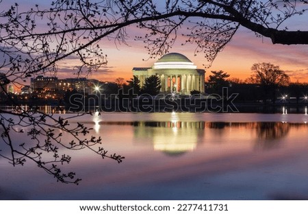 The Jefferson Memorial, a landmark framed by branches of a cherry tree about to blossom, reflecting in the water of the Potomac River along the Tidal Basin in downtown Washington, DC at sunrise.
