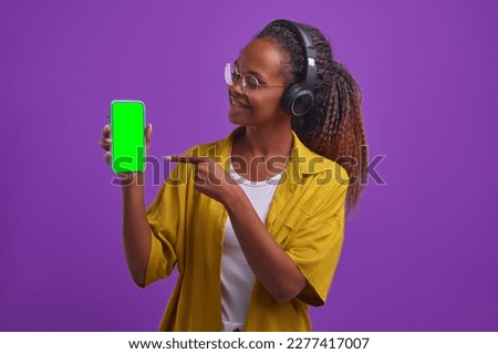 Young happy casual African American woman wearing headphones holding phone and pointing finger at green screen showing off buying flagship model of new smartphone or cool app stands in lilac studio