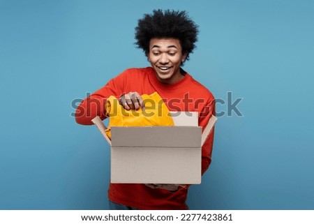 Young attractive overjoyed African American man influencer unpacking box isolated on blue background. Happy emotional guy with curly hair buying new clothes. Delivery, online shopping concept  Royalty-Free Stock Photo #2277423861