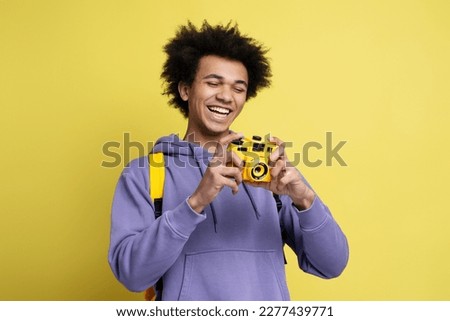 Happy curly haired guy photographer, journalist or traveler with yellow backpack, holding digital camera and smiling looking at camera, before taking photos isolated on yellow background 