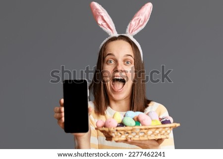 Photo of surprised amazed Caucasian woman wearing rabbit ears holding colorful Easter eggs and showing mobile phone with black empty display isolated on gray background. Copy space for advertisement.