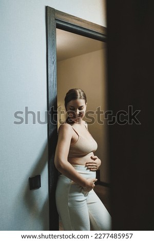 Cute young pregnant woman in a top and casual maternity clothes. Cute pregnant woman hugging her belly at home