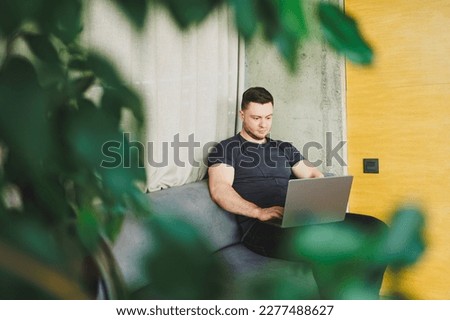 Smiling man in t-shirt sitting on sofa, typing on netbook, working remotely on startup as freelancer, looking at laptop and smiling