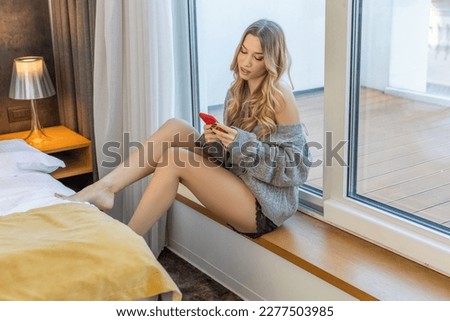 Beautiful young woman in panties and sweater using her smarthphone in hotel bedroom