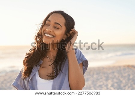 Smiling latin hispanic woman relaxing on beach with closed eyes at sunset. Beautiful mixed race woman enjoying wind fluttering hair. Charming young woman breathing fresh air at summer beach. Royalty-Free Stock Photo #2277506983