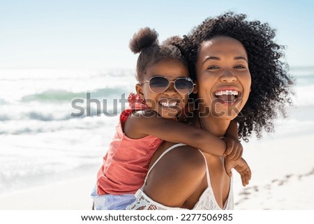 Smiling black mother and beautiful daughter wearing sunglasses having fun on the beach. Portrait of happy african american woman giving a piggyback ride to her cute little girl wearing shades. Royalty-Free Stock Photo #2277506985