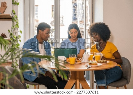 Multiethnic friends doing online shopping with the computer while having breakfast, by the window