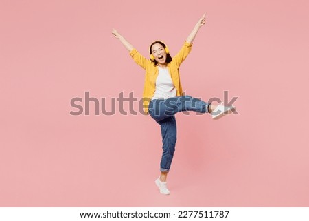 Full body young woman of Asian ethnicity wear syellow shirt white t-shirt headphones listen to music raise up leg spread hands dancing isolated on plain pastel light pink background studio portrait