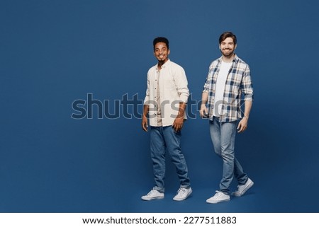 Full body side view young two friends happy men 20s wear white casual shirts together looking camera walking going strolling isolated plain dark royal navy blue background. People lifestyle concept