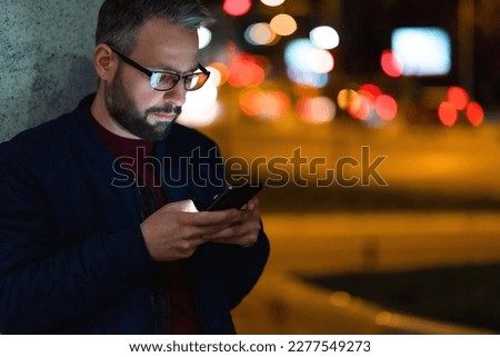 A man with glasses is using a smartphone in the city with a nightlight in the background.