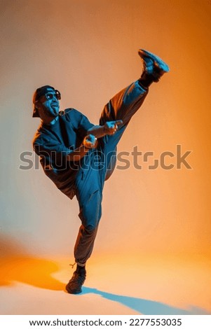 Cool stylish fun handsome young dancer man with fashion sunglasses and a cap in fashionable outfit with a t-shirt is dancing in a creative color studio with orange warm and neon cold light Royalty-Free Stock Photo #2277553035