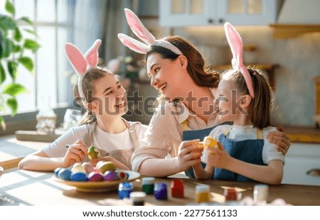 Happy mother and her daughters wearing aprons holding painted colorful eggs while decorating them with food dyes in cozy kitchen at home. Easter craft activities for families.