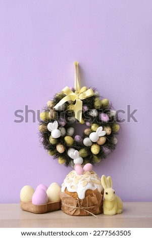 Table with Easter cake, eggs, rabbit and wreath on lilac wall