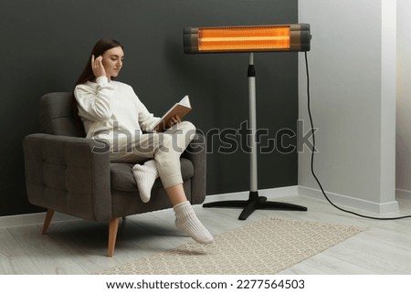 Young woman reading book on armchair near electric infrared heater at home. Space for text Royalty-Free Stock Photo #2277564503