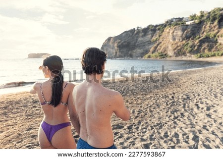 Handsome man wearing swimsuit with beautiful woman walks on the beach in a sunny day, holding hands, during holidays. Young adults caucasian couple travel together a romantic vacation relaxing outdoor