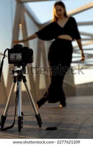 Vertical image of a young girl dancing on a pedestrian bridge and filming herself with a professional camera on a tripod on the ground.