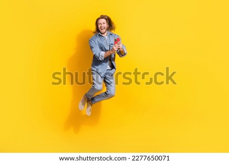 Full length photo of funny excited man wear denim jacket jumping high typing modern device isolated yellow color background