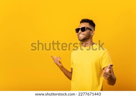 Excited positive young arab man in sunglasses rejoicing, raising hands up and having fun, having good summer mood. Indoor studio shot isolated on yellow background