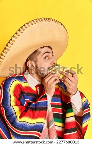Portrait of young man in colorful festive clothes, poncho and sombrero posing, eating taco against yellow studio background. Concept of mexican traditions, fun, celebration, festival, emotions