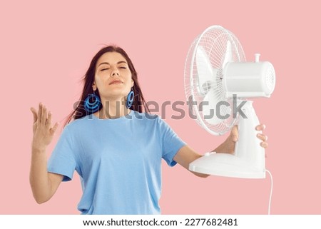 Ahh what a relief. Beautiful woman uses electric fan to survive summer heat and enjoys cool air on very hot days. Happy girl standing on pink studio background, holding fan and breathing fresh air