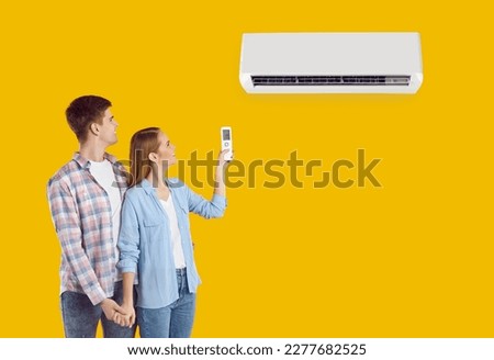Young couple switch air conditioner isolated on solid yellow color background. Happy millennial husband and wife using remote control to adjust AC unit temperature settings. Air conditioning concept