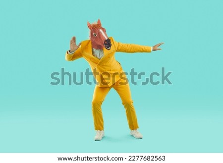 Funny man in a funny horse mask and a bright yellow suit is fooling around as if he is eavesdropping on someone. Full-size photo of a crazy showman on an isolated turquoise background.