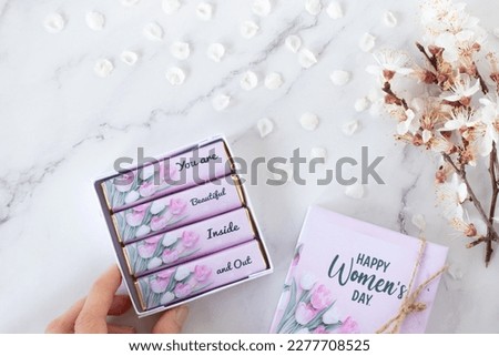 Happy Women's Day text on chocolate gift box with inspiring message and beautiful spring flower branch on white marble background. Top table view.
