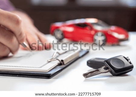 Man signing car insurance document or lease paper. Writing signature on contract or agreement. Buying or selling new or used vehicle. Car keys on table on red car background. Warranty or guarantee. Royalty-Free Stock Photo #2277715709