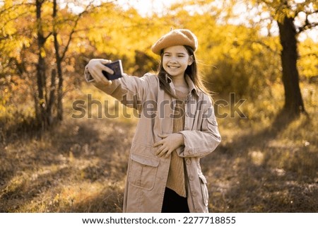 Young smiling girl walking in autumn park taking selfie pictures using smartphone, wearing beige coat and beret, happy mood, fashion style trend. Copy space