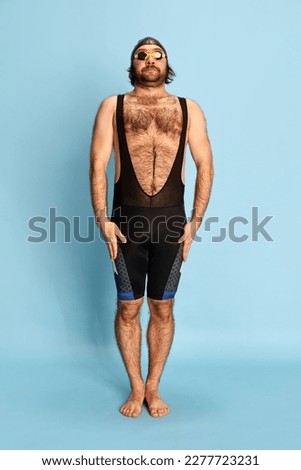 Comic portrait of funny bearded man in swimsuit and swimming cap and goggles having fun isolated over blue background. Concept of sport, active lifestyle and humor, funny meme emotions