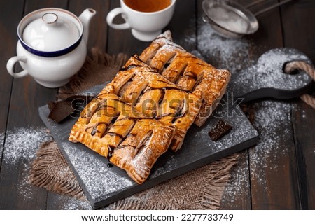 Homemade sweet puff pastries. Baked Puff pastry turnovers with chocolate and fruit filling served on a wooden board with cup and teapot on background. Selective focus, horizontal. Royalty-Free Stock Photo #2277733749