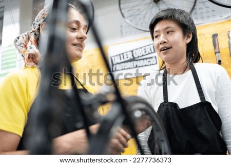 Two women in bike shop talking, a middle-aged Caucasian woman and a young Asian girl, both dressed in T-shirts and black aprons. One teaches the other bike maintenance in the workshop.