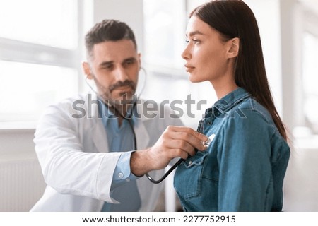 Lady coming to clinic for heart and lungs checkup, male doctor using stethoscope, listening to female patient's breath or heartbeat, sitting in clinic office Royalty-Free Stock Photo #2277752915