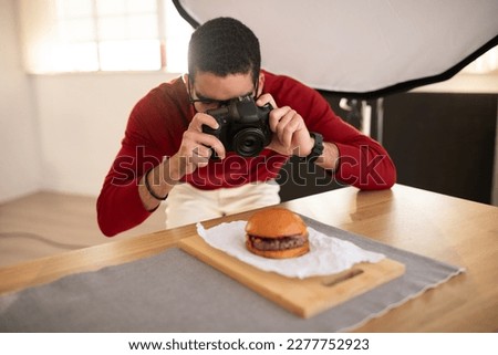 Food photographer handsome young middle eastern man wearing casual outfit and eyeglasses taking photos of delicious juicy meaty hamburger at photo studio, using dslr camera