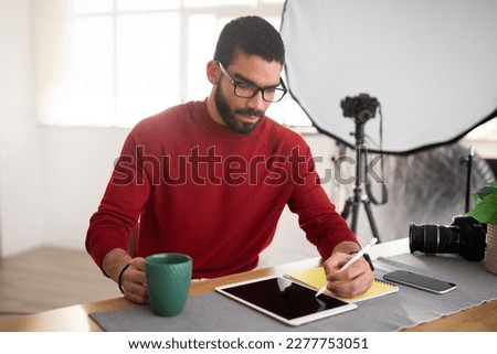 Handsome bearded millennial hispanic man photographer wearing eyeglasses sitting at table, working on pc digital pad with black empty screen at office studio, using stylus pencil, drinking coffee