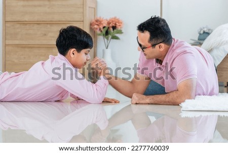 Indian happy father and son playing together, laying down on floor, doing arm wrestle with amusement and fun while staying in living room at cozy home in weekend. Education, Family, Activity Concept