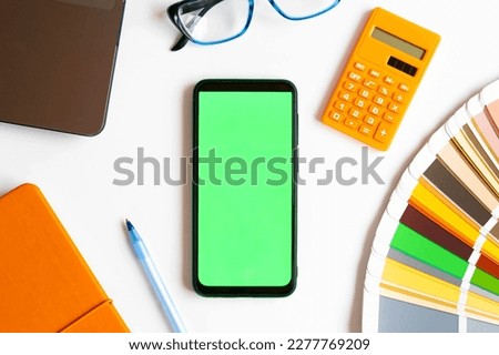 Top view of Mobile Phone Device Green Screen on Designer Worktop. Close Up. Color Guide, Palette, Laptop, Pen. Cell Phone Mockup with Blank Chromakey Display on Workplace. Smartphone Layout Color key