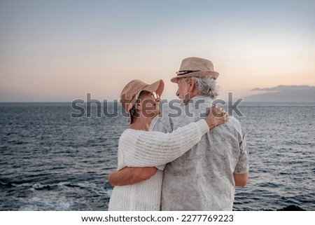 Back view of romantic senior couple or pensioners embraced at the sea at sunset light expressing love and tenderness - old couple outdoors enjoying vacations together