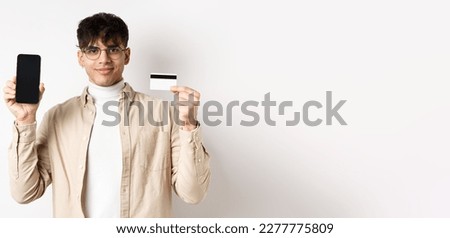 Online shopping. Young modern guy showing plastic credit card and empty smartphone screen, demonstrate account, standing on white background.